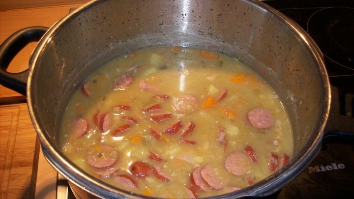 In einem gro&szlig;en Topf k&ouml;chelt eine hausgemachte Erbsensuppe mit Wurstscheiben und buntem Gem&uuml;se auf einem Herd.
