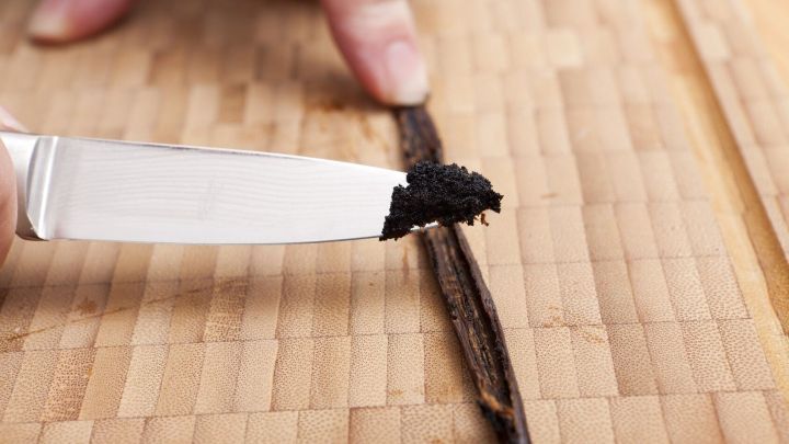 Eine Hand schneidet eine Vanilleschote auf einem Holztisch, w&auml;hrend das schwarze Vanillemark auf das Messer gerieben wird.
