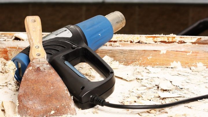 Ein Hei&szlig;luftgebl&auml;se und ein Spachtel liegen auf einer Holzoberfl&auml;che, w&auml;hrend Lackschichten abgetragen werden.