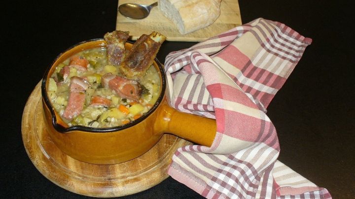 Ein rustikaler Tontopf mit bunter Graupensuppe und knusprigen Rippchen steht auf einem Holzbrett, umgeben von einem karierten Tuch.