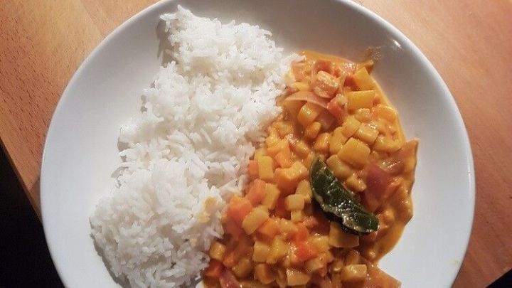 Ein Teller mit asiatischem Gem&uuml;se-Curry liegt neben einer Portion Reis auf einem Holztisch. Das Curry ist farbenfroh und enth&auml;lt verschiedene Gem&uuml;sesorten.