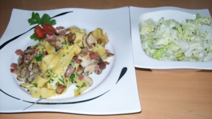Eine bunte Schinken-Pfanne mit frischen Champignons und Nudeln liegt auf einem wei&szlig;en Teller neben einem Salat.