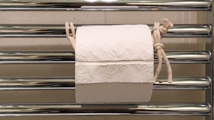 Ein Toilettenpapier-Halter h&auml;lt eine Rolle in einem kleinen Bad, umgeben von modernen Fliesen und gl&auml;nzenden Stangen.
