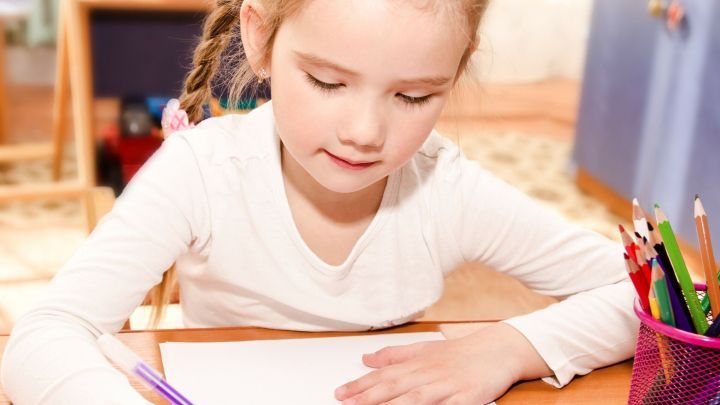 Ein Kind sitzt an einem Tisch und schreibt mit einem Buntstift auf ein leeres Blatt Papier, um Vorschul&uuml;bungen zu machen.
