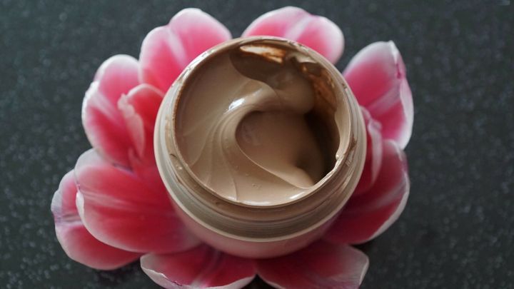 Eine runde, offene Salbenschachtel mit cremiger Textur liegt auf einer pinken Bl&uuml;te, umgeben von einer dunklen Oberfl&auml;che.