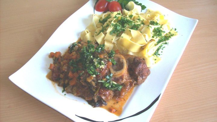 Ein Teller Ossobuco ist mit frischer Gremolata garniert. Daneben liegen Pasta und ein frischer Salat.