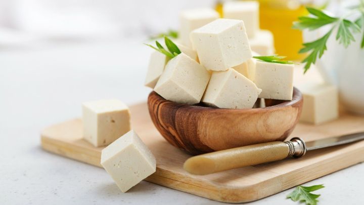 W&uuml;rfel aus Tofu liegen in einer Holzschale, umgeben von frischen Kr&auml;utern auf einem Holzbrett, perfekt f&uuml;r die Zubereitung.