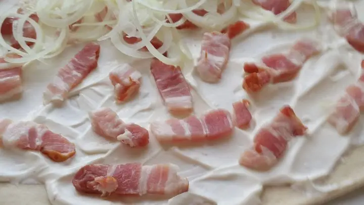 Den klein geschnittenen Speck und auch die Zwiebelringe auf der Schmand-Creme-fraiche-Masse verteilen.