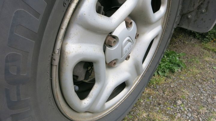 Ein Alufelgen-Rad steht auf einem unebenen Untergrund, sichtbar mit Schmutz und Gras. Die Felge ist leicht abgenutzt.