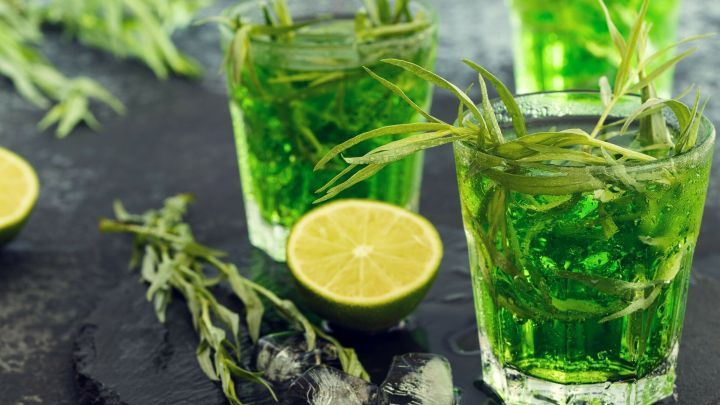 Gr&uuml;ne, erfrischende Estragon-Getr&auml;nke stehen in Gl&auml;sern, umgeben von Limetten und Estragon, auf einem dunklen Untergrund.