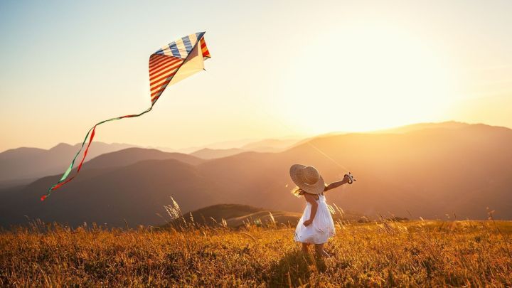 Ein Kind spielt fr&ouml;hlich im Sonnenlicht und l&auml;sst einen Drachen in der warmen Fr&uuml;hlingsluft fliegen, umgeben von Bergen.