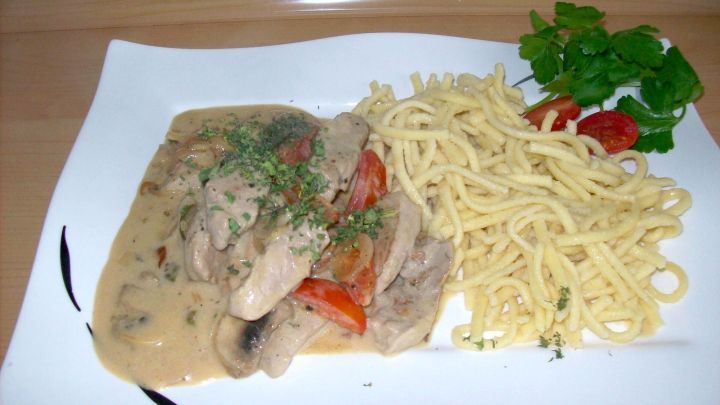 Geschmackvolles Schweinefilet &agrave; la Stroganoff wird neben frischen Nudeln und Kr&auml;utern auf einem wei&szlig;en Teller serviert.