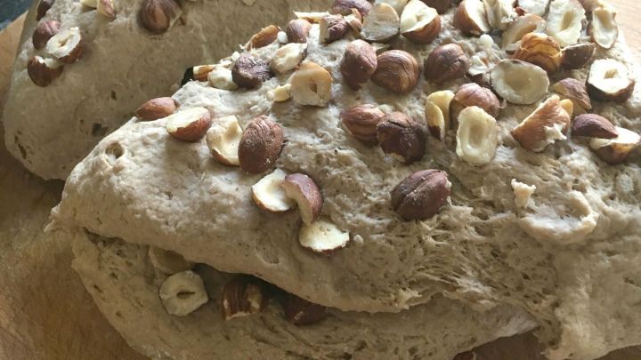 Zwei frisch gebackene Brote mit N&uuml;ssen liegen auf einem Holzbrett, w&auml;hrend eine Hand das eine Brot greift.