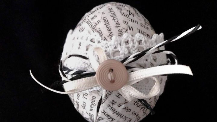 Ein dekoriertes, ausgeblasenes Ei im Zeitungslook liegt auf einem schwarzen Hintergrund, geschm&uuml;ckt mit B&auml;ndern und einem Knopf.