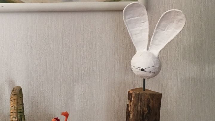 Ein wei&szlig;er Osterhase aus Papier sitzt auf einem Holzst&uuml;ck in einem gem&uuml;tlichen Wohnraum neben einem geflochtenen Korb.
