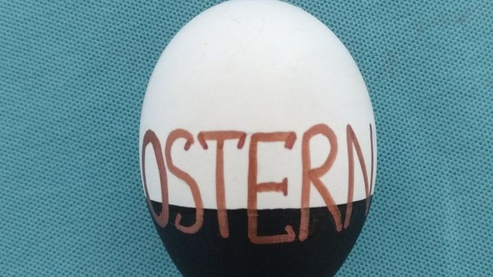 Ein Ei, mit Acrylfarbe in Schwarz und Wei&szlig; gestaltet, zeigt das Wort "OSTERN" in braunen Buchstaben auf einer unifarbenen Fl&auml;che.