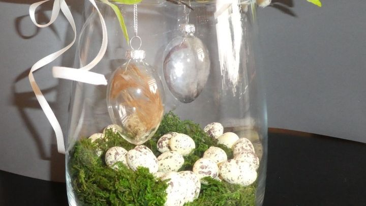 Ein Glasgef&auml;&szlig; mit Moos und kleinen Eiern, dekoriert mit modernen Ostereiern und einem Zweig, der frisch wirkt.