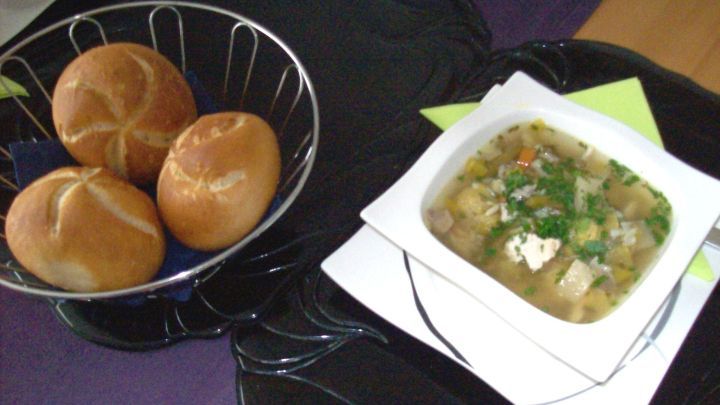 Eine Sch&uuml;ssel Gem&uuml;sesuppe mit frischem Gartengem&uuml;se steht neben einem Korb mit drei Br&ouml;tchen auf einem Tisch.