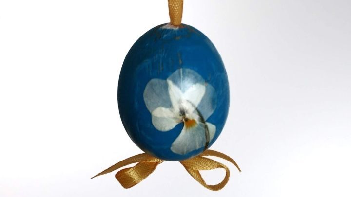 Ein blaues Osterei mit einer Bl&uuml;te h&auml;ngt dekorativ an einem gelben Band von einem Zweig, vor hellem Hintergrund.