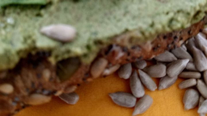 Ein gr&uuml;ner Brotaufstrich mit Rucola und Sonnenblumenkernen liegt auf einem Teller, umgeben von einigen Sonnenblumenkernen.