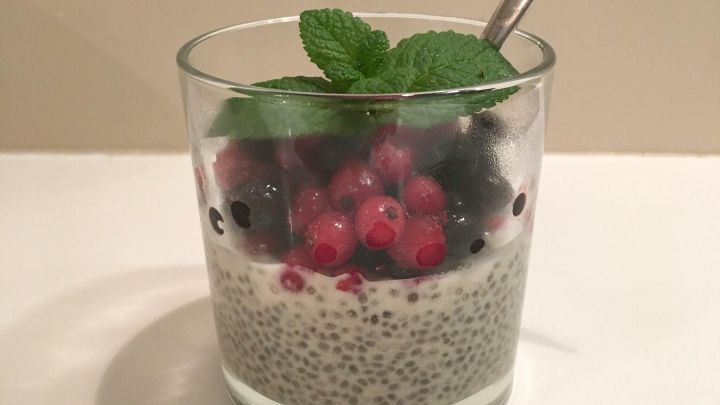 Ein Glas mit Chia-Samen-Pudding, garniert mit frischen Beeren und Minze, steht auf einem wei&szlig;en Tisch. Ein L&ouml;ffel ist darin.
