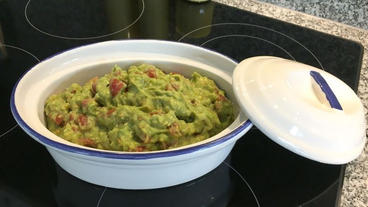 Ein gef&uuml;llter, wei&szlig;er Beh&auml;lter mit einem Avocado-Tomaten-Dip steht auf einer Herdplatte aus schwarze. Der Deckel ist teilweise ge&ouml;ffnet.
