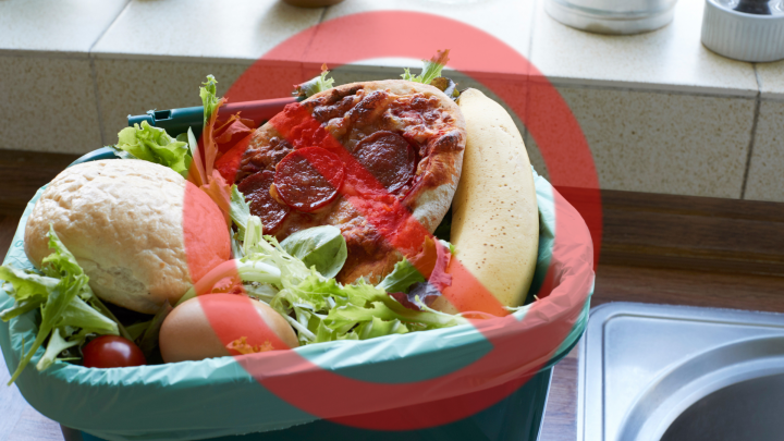 Ein &uuml;bervoller M&uuml;lleimer mit Lebensmitteln, darunter eine alte Pizza und Gem&uuml;se, steht in einer K&uuml;che.