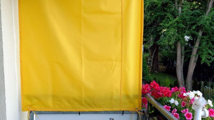 Ein gelbes Sonnenschutz-Paneel h&auml;ngt auf einem Balkon, um Schatten zu spenden, umgeben von bunten Blumen.