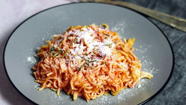 Ein Teller mit Tomatennudeln wird appetitlich angerichtet, umgeben von einem grauen Besteckset auf einem sanften Untergrund.
