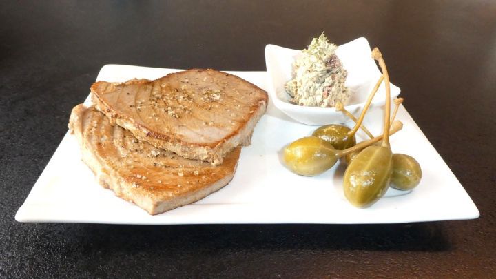 Gegrilltes Thunfischsteak liegt saftig auf einem wei&szlig;en Teller, begleitet von w&uuml;rziger Sardellenbutter und Kapernbeeren.