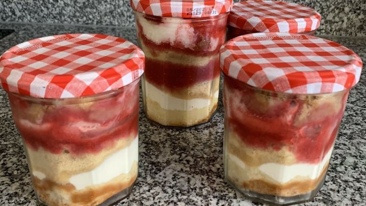 Vier Gl&auml;ser mit fruchtigem Erdbeerdessert stehen auf einer grauen Arbeitsplatte, gef&uuml;llt mit Schichten aus Erdbeeren, Sahne und Keksen.