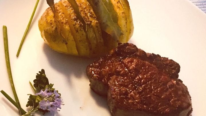 Eine ofenfrische schwedische Ofenkartoffel mit Lorbeerbl&auml;ttern liegt neben saftig gebratenem Fleisch, dekoriert mit Kr&auml;utern.