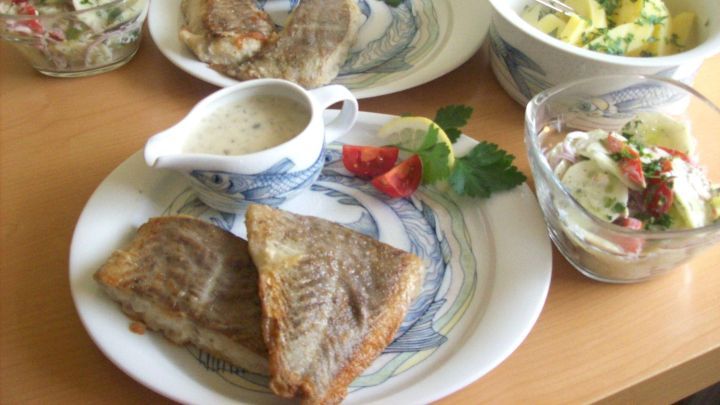 Gebratener Seelachs liegt auf Tellern, umgeben von Bechamel-So&szlig;e und frischem Gem&uuml;se in Sch&uuml;sseln auf einem Tisch.