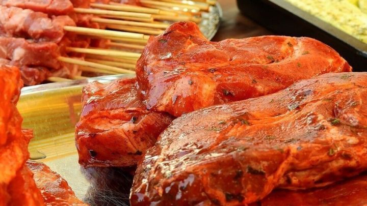 Saftiges Schweinefleisch, mariniert in w&uuml;rziger Mischung, liegt auf einem Tisch neben Grillspie&szlig;en und Kr&auml;utern.