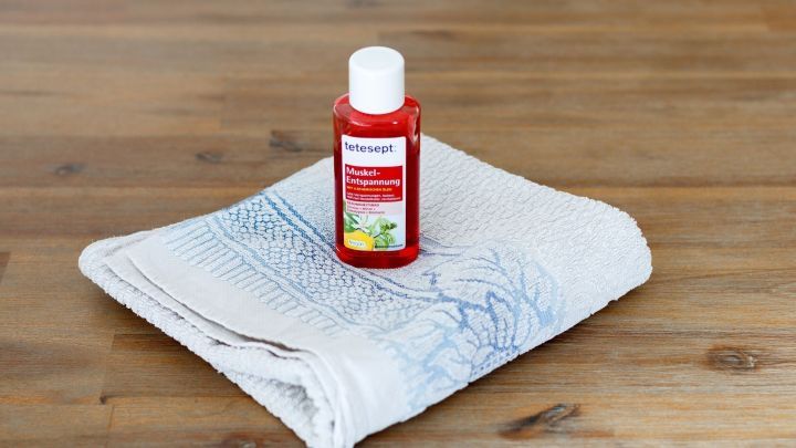 Eine rote Flasche mit Badezusatz steht auf einem wei&szlig;en Handtuch, bereit f&uuml;r ein warmes Bad gegen Muskelkater.