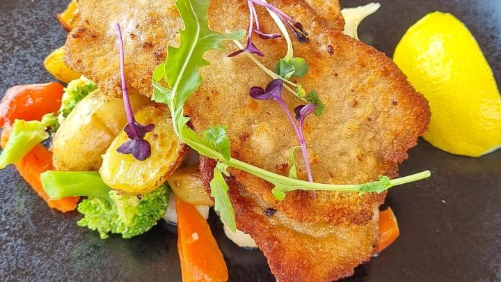 Panierter Fisch liegt auf einem schwarzen Teller, umgeben von buntem Gem&uuml;se und einer Zitronenscheibe.