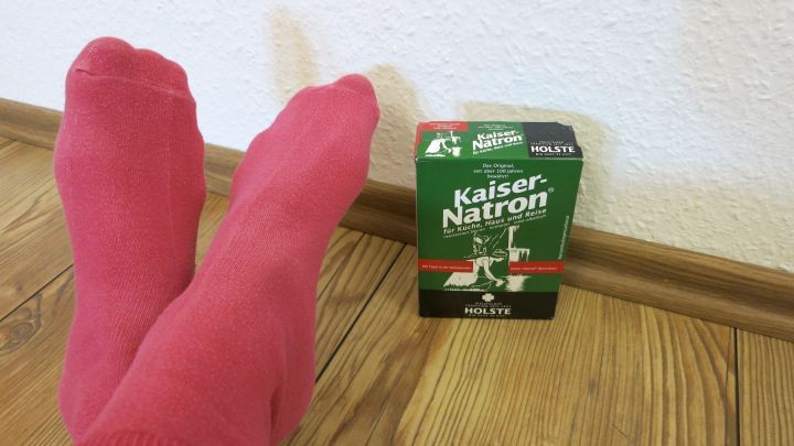 Rosa Socken liegen entspannt auf einem Holzboden neben einer gr&uuml;nen Natronverpackung, um Schwei&szlig;f&uuml;&szlig;e zu behandeln.