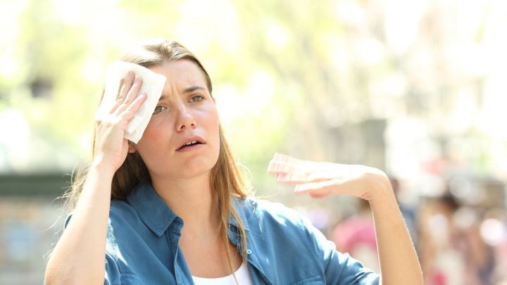 Eine Frau wischt sich den Schwei&szlig; von der Stirn, w&auml;hrend sie in einer warmen, sonnigen Umgebung im Freien steht.