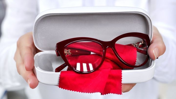 Ein rotes Kunststoff-Brillengestell liegt in einer wei&szlig;en Box, w&auml;hrend eine Hand es vorsichtig h&auml;lt, umgeben von einem roten Reinigungstuch.