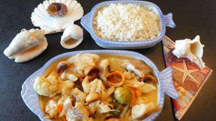 Ein schmackhaftes Fischragout mit Gem&uuml;se in einer Kokosso&szlig;e wird in einer blauen Fischform serviert, begleitet von Reis und Muscheln.