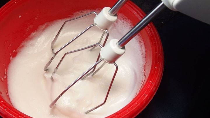 Eine R&uuml;hrmaschine schl&auml;gt Eiwei&szlig; in einer roten Sch&uuml;ssel, die auf einer dunklen Arbeitsfl&auml;che steht.