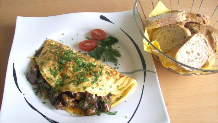 Ein Omelette Chasseur liegt auf einem wei&szlig;en Teller, garniert mit Kr&auml;utern und Tomatenscheiben, neben einem Korb mit Brot.