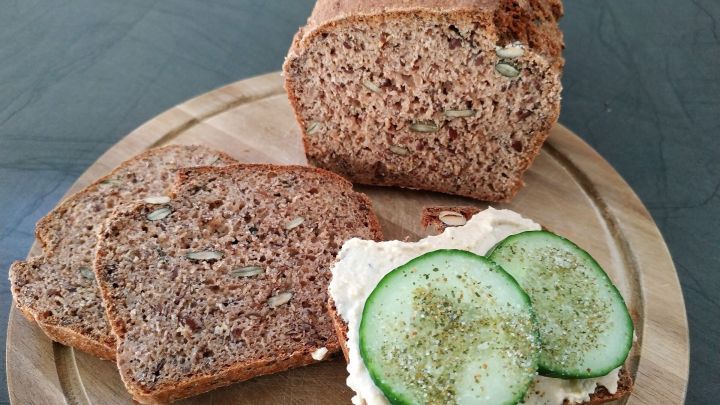 Ein freistehendes Vollkornbrot liegt auf einem Holzbrett, neben einigen geschnittenen Scheiben und einer belegten Scheibe mit Gurken.