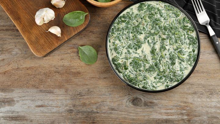 Ein cremiger Rahmspinat in einer schwarzen Sch&uuml;ssel liegt neben frischem Knoblauch und gr&uuml;nem Blattspinat auf einem Holztisch.