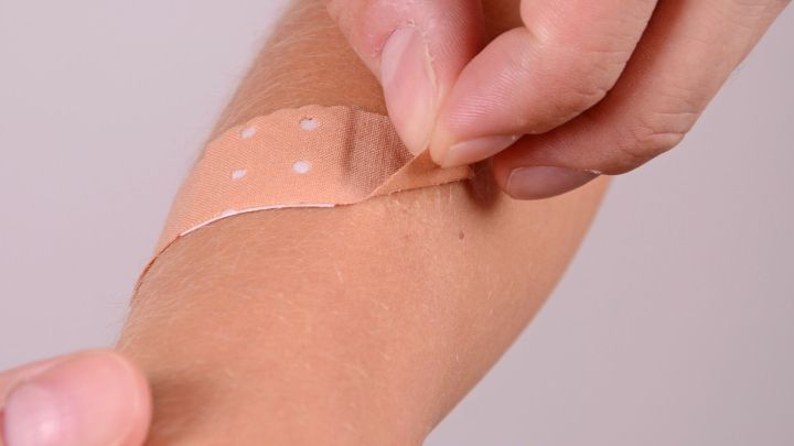 Ein Hand greift ein Wundpflaster, das sanft von einem Arm abgezogen wird, in einem neutralen, hellen Hintergrund.