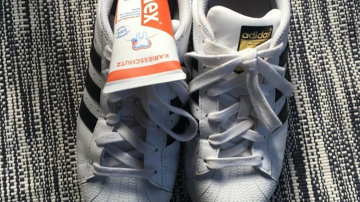 Wei&szlig;e Sportschuhe liegen auf einem gestreiften Teppich, w&auml;hrend ein Fleckenentferner daneben pr&auml;sentiert wird.