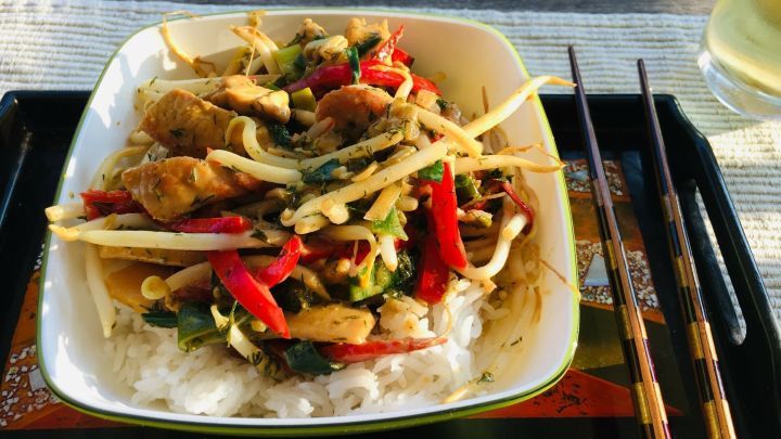 Ein Teller mit asiatischer Pute liegt auf einem Tisch, umgeben von frischem Gem&uuml;se, Reis und Essst&auml;bchen.