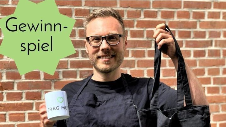 Eine Person h&auml;lt eine Tasse und eine Tasche in der Hand, l&auml;chelt und pr&auml;sentiert die Gewinne eines Gewinnspiels vor einer Backsteinwand.