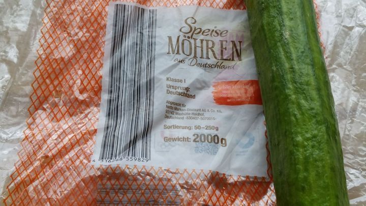 Ein langer gr&uuml;ner Gurke liegt neben einem transparenten Frischhaltebeutel f&uuml;r Karotten auf einem Tisch.