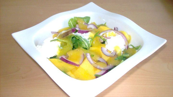 Ein frischer Rucola-Salat mit Mango und Zwiebeln in einer wei&szlig;en Schale auf einem Holzuntergrund.